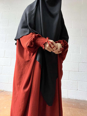 A close-up of a woman wearing a red Butterfly Abaya with smocked sleeves, her hands clasped in front, and a black headscarf.