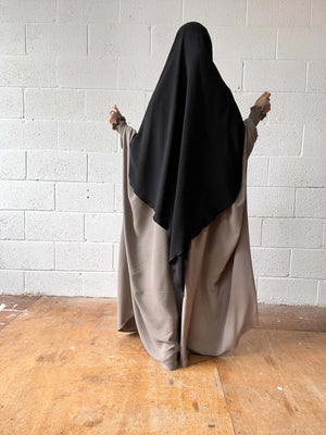 A woman in a taupe Butterfly Abaya with a black headscarf standing against a white brick wall, photographed from the back.