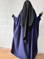 A woman wearing a navy blue Butterfly Abaya with a black headscarf standing against a white brick wall, photographed from the back.