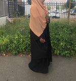 Woman wearing a long beige hijab over a black robe, standing outside by a fence, with the hijab flowing gently.