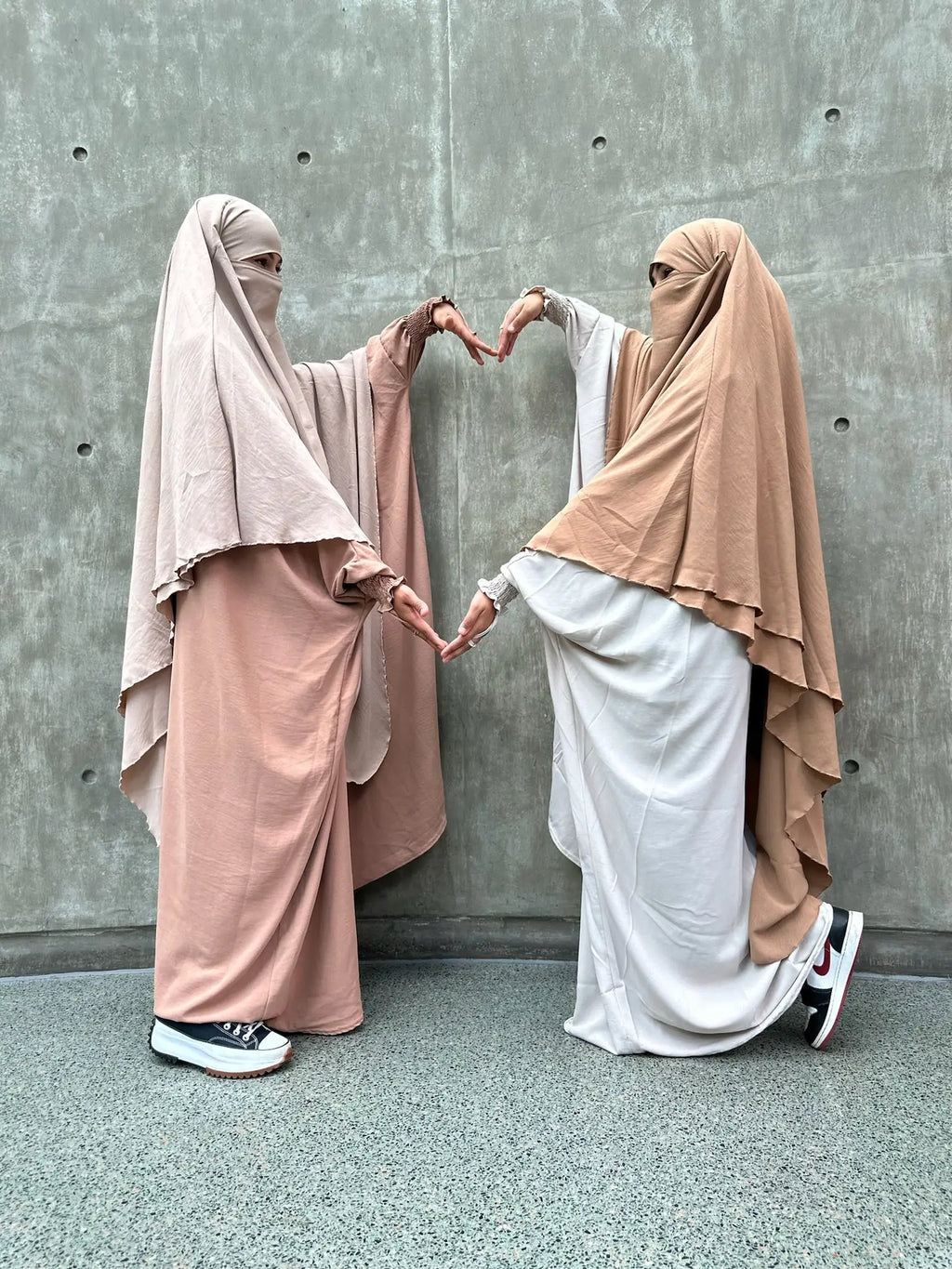 Two women in beige and taupe Butterfly Abayas creating a heart shape with their hands in front of a concrete wall.