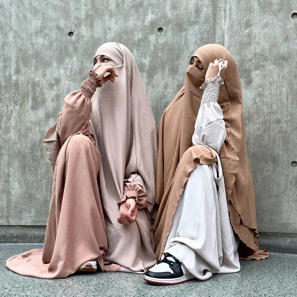 Two women wearing beige and taupe Butterfly Abayas sitting against a concrete wall, with soft fabric details.