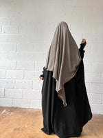 A woman in a black Butterfly Abaya with a beige headscarf, standing against a white brick wall, photographed from behind.