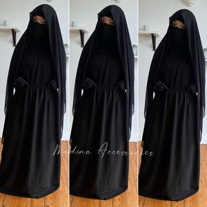 Modest fashion outfit featuring a woman in a dark black square hijab and a black dress, standing in a modern living room.