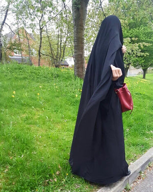 Side view of a woman in a park, wearing a long black hijab and carrying a red bag, with trees in full bloom behind her.