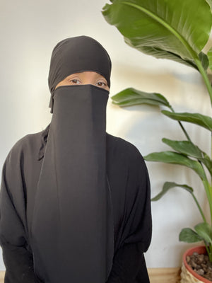 Woman in a black niqab seated in front of tropical plants, looking pensively towards the camera.