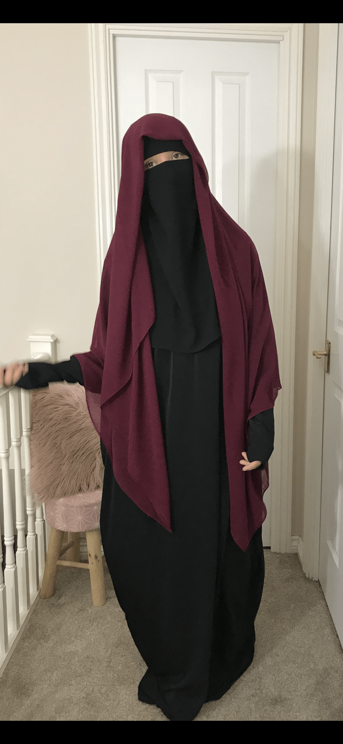 Woman in a dark home setting adjusting a deep burgundy long hijab over a niqab, the fabric flowing down to her waist.