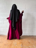 A woman in a maroon Butterfly Abaya, standing against a white brick wall, photographed from behind.