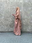 A woman in a beige Butterfly Abaya standing against a concrete wall, shown from the side.