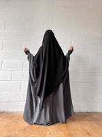 A woman in a dark grey Butterfly Abaya standing against a white brick wall, shown from the back with arms slightly raised.