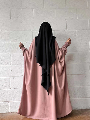 A woman in a pink Butterfly Abaya and black headscarf standing against a white brick wall, photographed from the back.