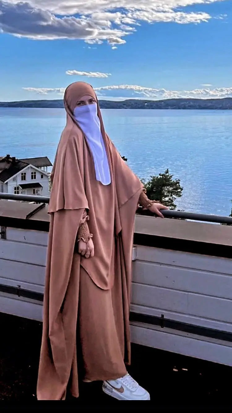 A woman wearing a beige Butterfly Abaya and white face veil, standing by a waterfront railing with a scenic lake and sky in the background.