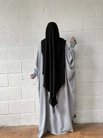 A woman in a grey Butterfly Abaya with a black headscarf standing against a white brick wall, photographed from the back.
