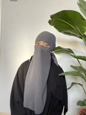 Woman wearing a light gray niqab, partially covering her face with her hand, standing indoors beside a large green plant.