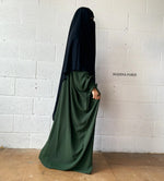 A woman in a green Butterfly Abaya and black headscarf standing against a white brick wall, captured in a side profile.