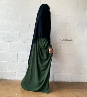 A woman in a green Butterfly Abaya and black headscarf standing against a white brick wall, captured in a side profile.