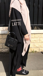 Woman outside, covered in a light creme square hijab over a black robe, standing by a metal fence, the fabric draped gracefully.