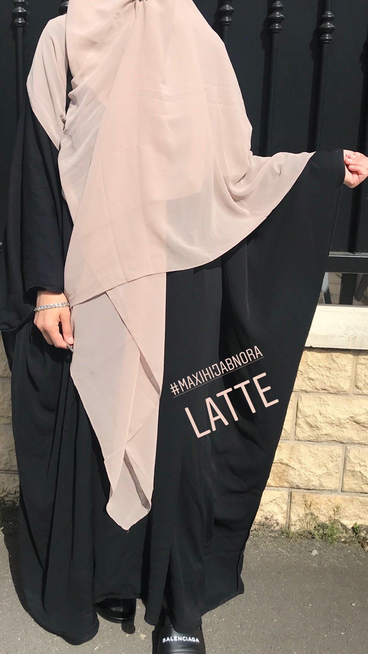 Woman styled in a light pink square hijab labeled 'Latte', draped over a black robe, posing outside against a stone wall.