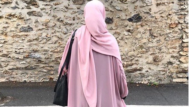 Rear view of a woman outdoors, wearing a pastel pink long hijab over a purple robe, surrounded by lush greenery.