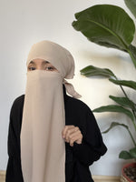 Woman covered with a beige niqab, indoors, with holding it, standing beside a green potted plant.