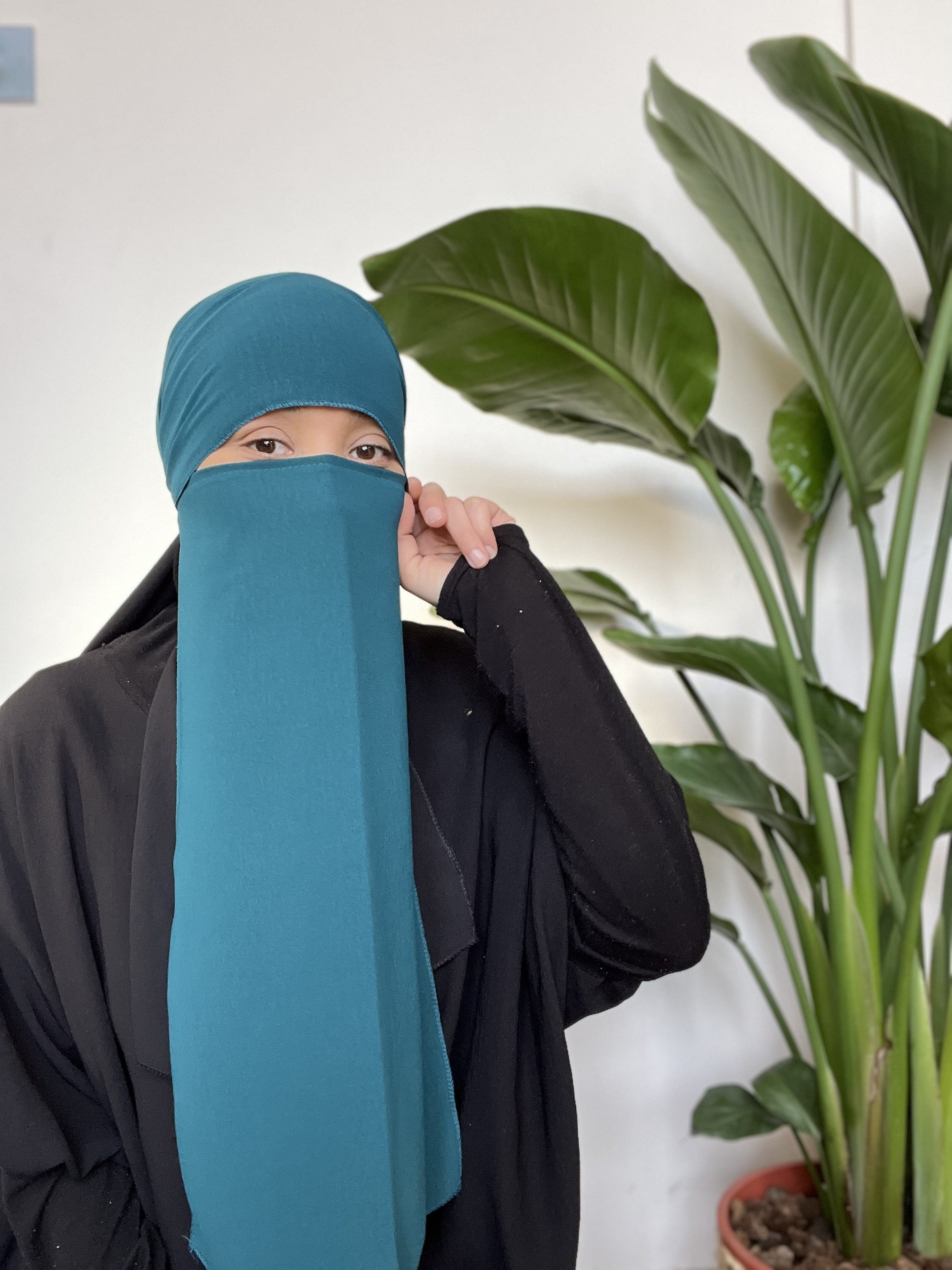 Woman in a teal niqab partially covering her face, with large indoor plants in the background.