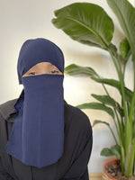 Woman wearing a dark navy niqab covering her face, standing in a bright indoor setting with tropical plants.