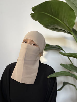 Woman wearing a light beige niqab covering her face, standing in front of green indoor foliage.