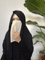 Woman in cream niqab looking at the camera with a serene expression, large green leaves in the background.