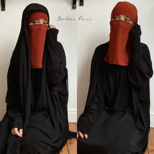 Dual images of a woman in a niqab, one red and one black, sitting on the floor in a minimalist decor.