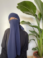 Woman in a deep blue niqab covering her face, standing beside green indoor plants.