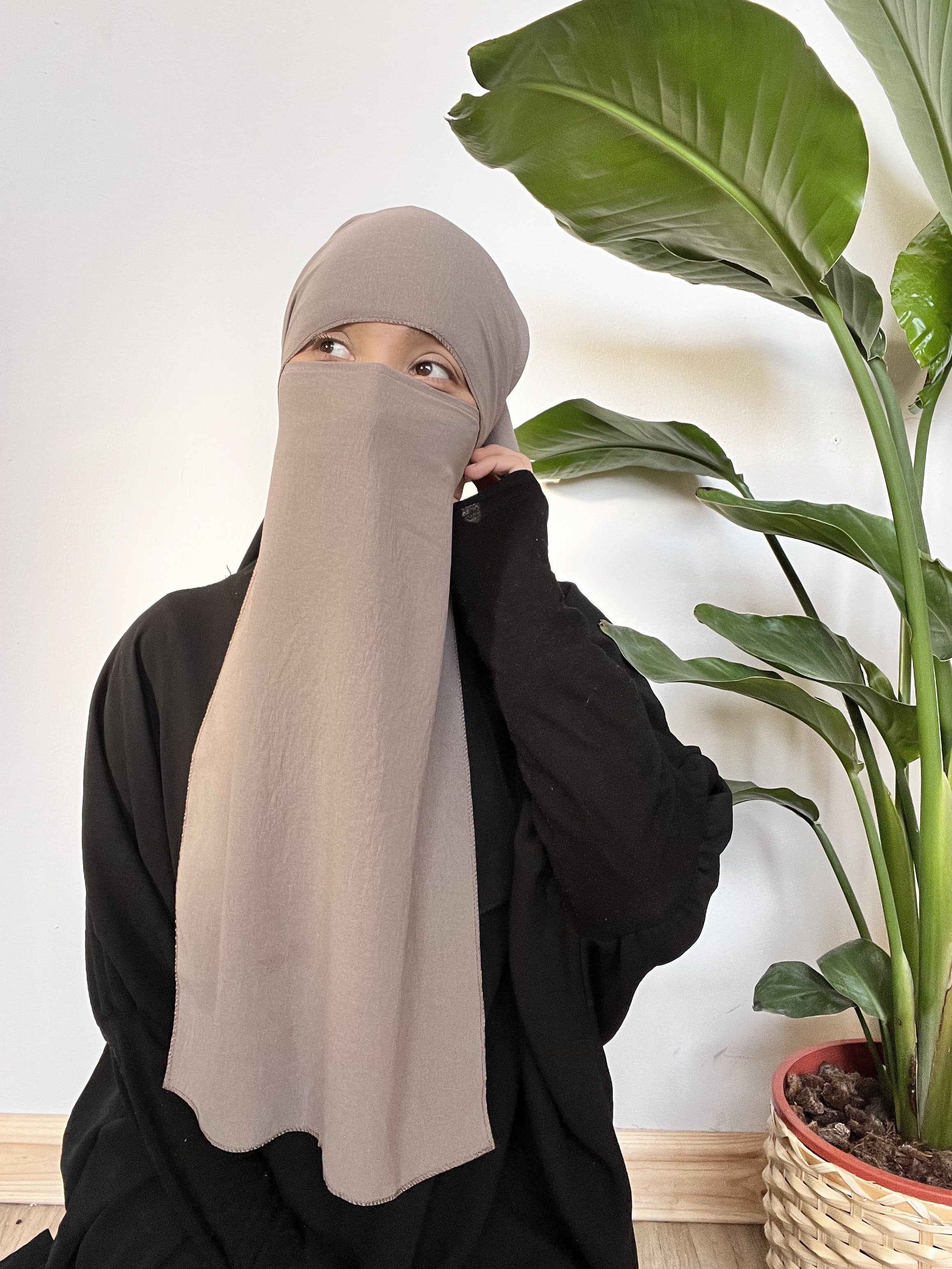 Woman wearing a light gray niqab, partially covering her face with her hand, standing indoors beside a large green plant.