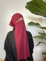 Woman in a bright maroon niqab covering her face, standing in front of a large leafy plant in an indoor setting.