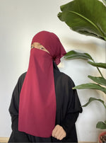 Woman in a maroon niqab posing with her hand on her cheek, standing in front of lush greenery indoors.