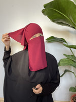 Woman in a maroon niqab gesturing upwards, seated on the floor in a casual indoor environment.