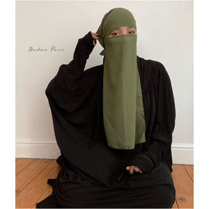 Woman in an olive green niqab posing playfully while sitting on the floor in a casual setting