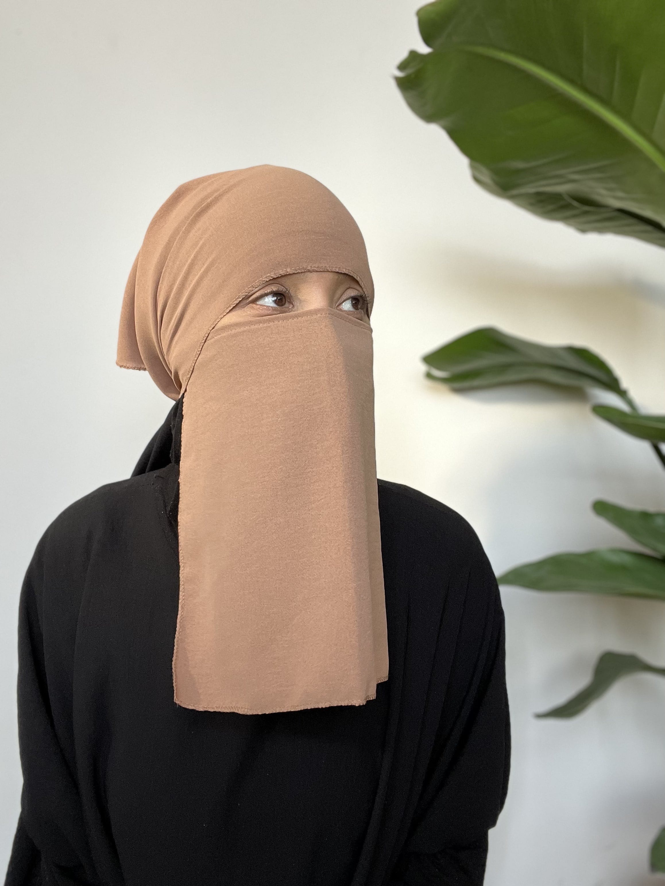 Woman covered with a sand-colored niqab, standing in an indoor setting adorned with green plants.