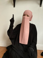 Woman in a pink niqab gesturing upwards, seated on the floor in a casual indoor environment.