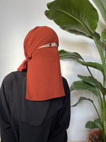 Woman in a rust-red niqab looking sideways, set against a background of lush indoor greenery.