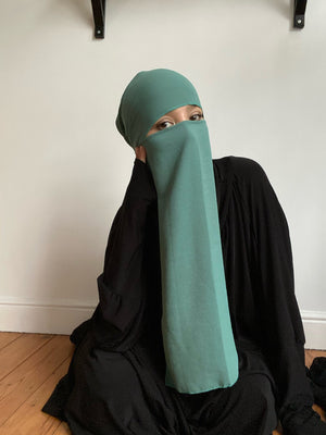 Woman in teal niqab seated on the floor in a minimalist room, looking thoughtfully to the side.