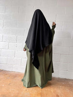 A woman in an olive green Butterfly Abaya and black headscarf standing against a white brick wall, shown from behind.