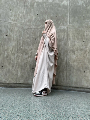 A woman in a beige and white layered Butterfly Abaya with a matching face veil, standing in front of a concrete wall, wearing black and white Nike sneakers.