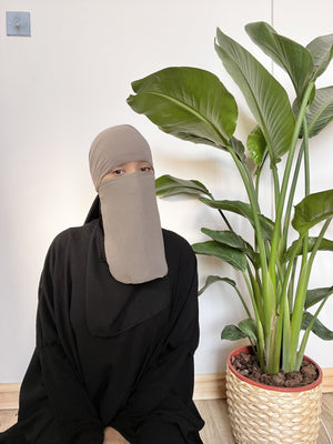 Woman in a light brown niqab seated in front of tropical plants, looking pensively towards the camera.