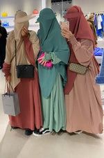 Three women dressed in modest Butterfly Abayas in muted tones (pink, green, beige) taking a selfie in a store, each holding designer handbags.