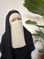 Woman wearing a cream niqab in a modern indoor setting with a lush green plant backdrop.