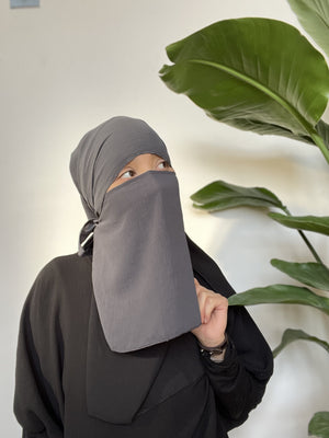 Woman in a grey niqab standing indoors, covering her face completely, beside vibrant green foliage.