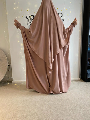 A woman in a dusty pink Butterfly Abaya standing in front of a decorated wall with string lights, shown from the back.