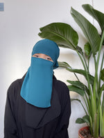 Woman in a turquoise niqab with only her eyes visible, in a serene indoor setting with lush greenery.