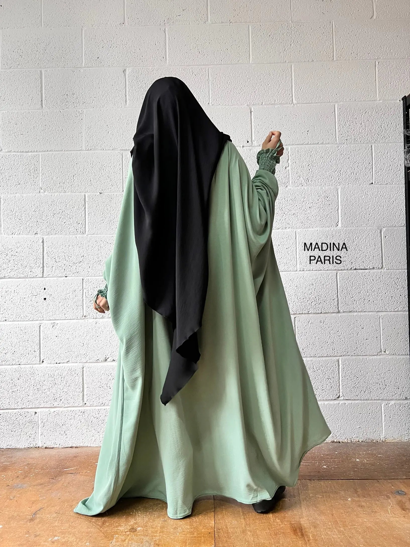 A woman wearing a mint green Butterfly Abaya with a black headscarf standing against a white brick wall, photographed from the back.