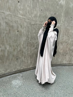 A woman in a white Butterfly Abaya with black accents adjusting her face veil, standing in front of a textured concrete wall.